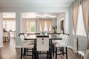 Un espace repas avec une longue table et de nombreuses chaises, une cuisine à l’arrière-plan et une vue sur l’extérieur grâce à de grandes fenêtres.