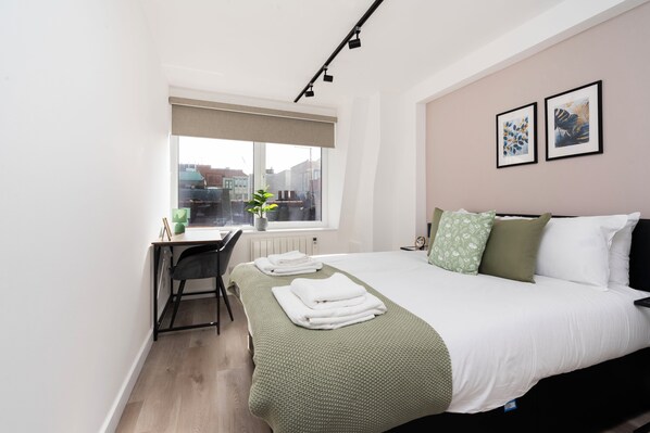 A bedroom with a bed, a desk, a chair, a radiator, and a window with a view of buildings.