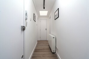 A narrow hallway with white walls, a wooden floor, a ceiling light, a radiator, and a door at the end.