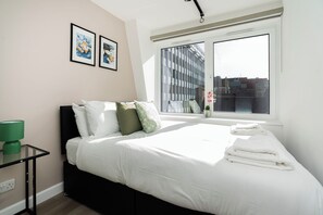 Un lit bien fait, avec des oreillers, une table de chevet avec une lampe et une fenêtre donnant sur des immeubles.