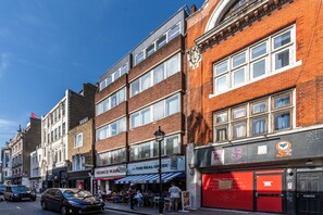 Un immeuble à plusieurs étages dont le rez-de-chaussée abrite divers commerces, notamment une boulangerie et un restaurant grec.