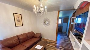 Un salon avec un canapé en cuir marron, une table basse en bois, un lustre, un tableau encadré et un téléviseur.