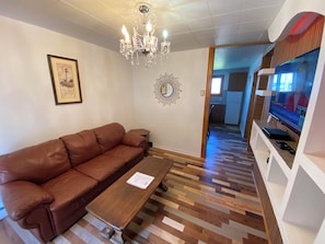 Una sala de estar con un sofá de cuero marrón, una mesa de centro de madera, una araña, un cuadro enmarcado y un televisor.