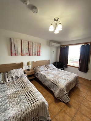 Habitación de hotel con dos camas, ventilador de techo, aire acondicionado, ventana con cortinas y una obra de arte colgada en la pared.