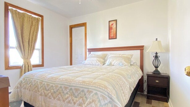 Une chambre à coucher comprenant un lit, une table de chevet, une lampe, un ventilateur de plafond et une fenêtre avec des rideaux.