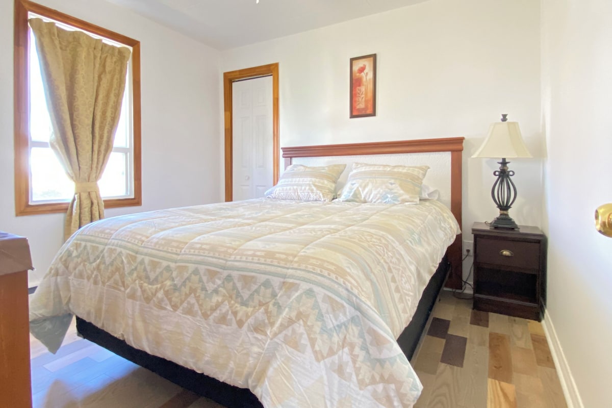 Un dormitorio con cama, mesita de noche, lámpara, ventilador de techo y ventana con cortinas.