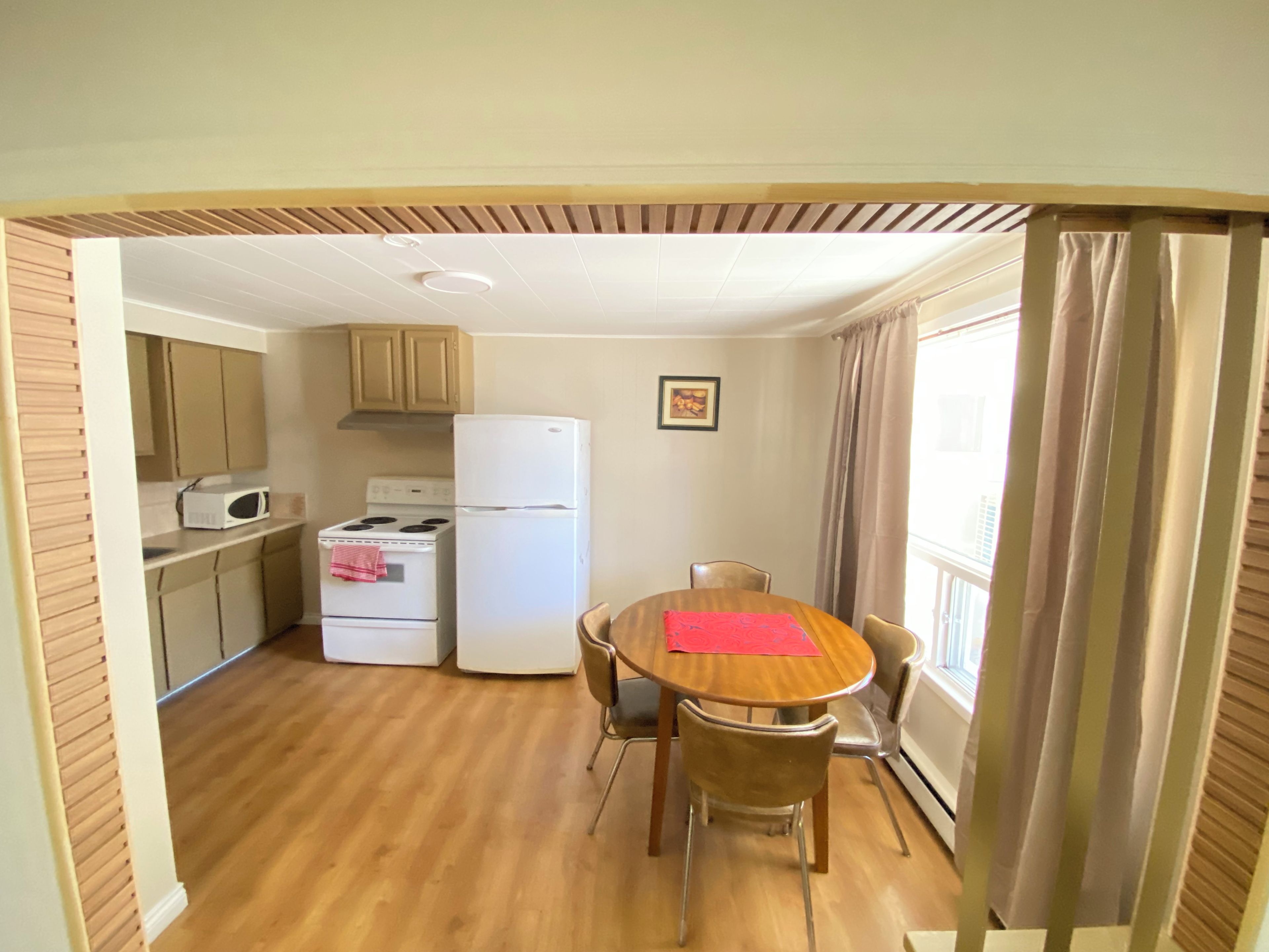 A compact kitchen with a microwave, stove, and refrigerator. There is a dining table with chairs and a window with curtains.