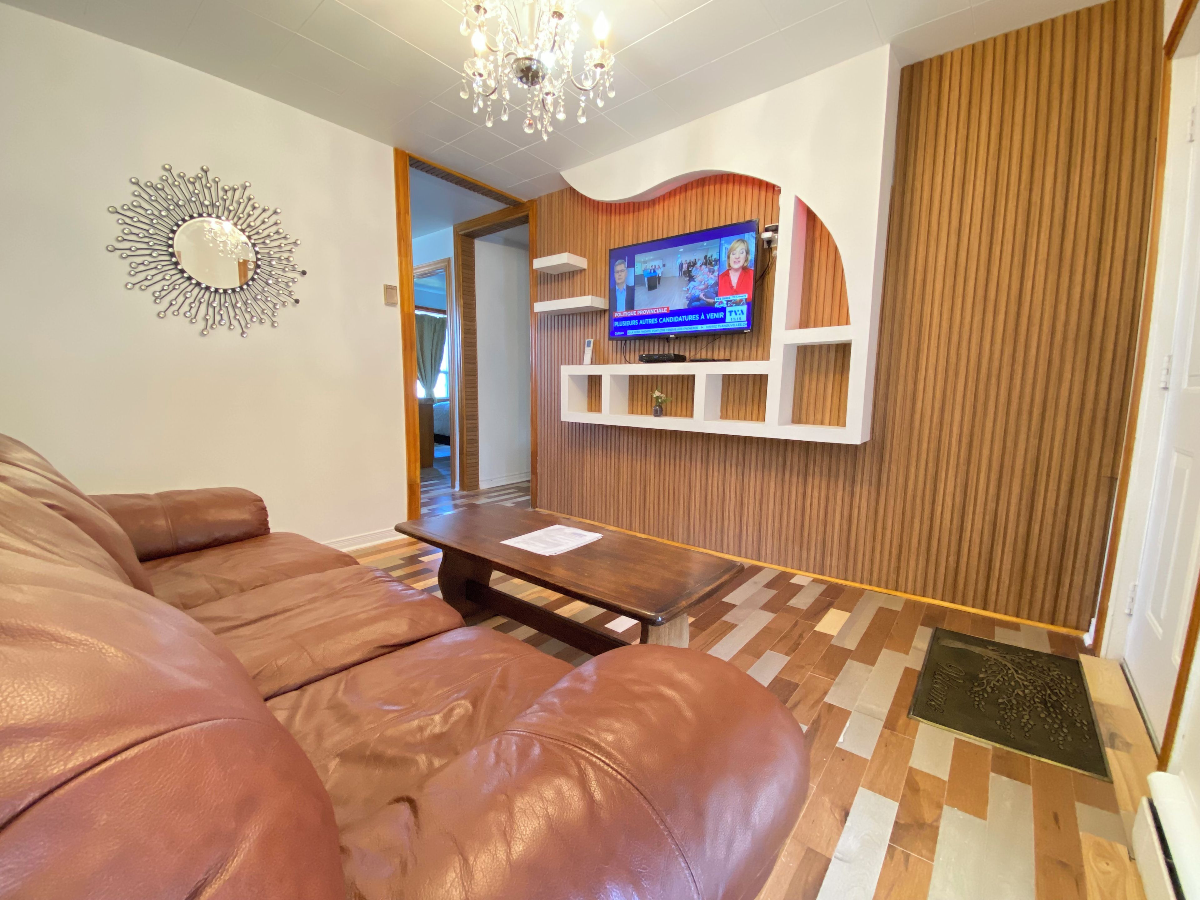 A living room with a leather sofa, a wooden coffee table, a television mounted on a wooden wall, and a chandelier.