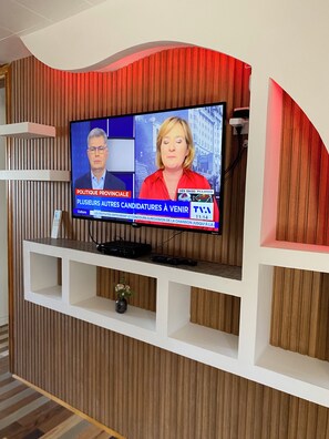 A television mounted on a wall with a wooden panel design, a white shelf below, and a remote control on the shelf.