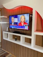 Un televisor montado en una pared con diseño de panel de madera, un estante blanco debajo y un control remoto sobre el estante.
