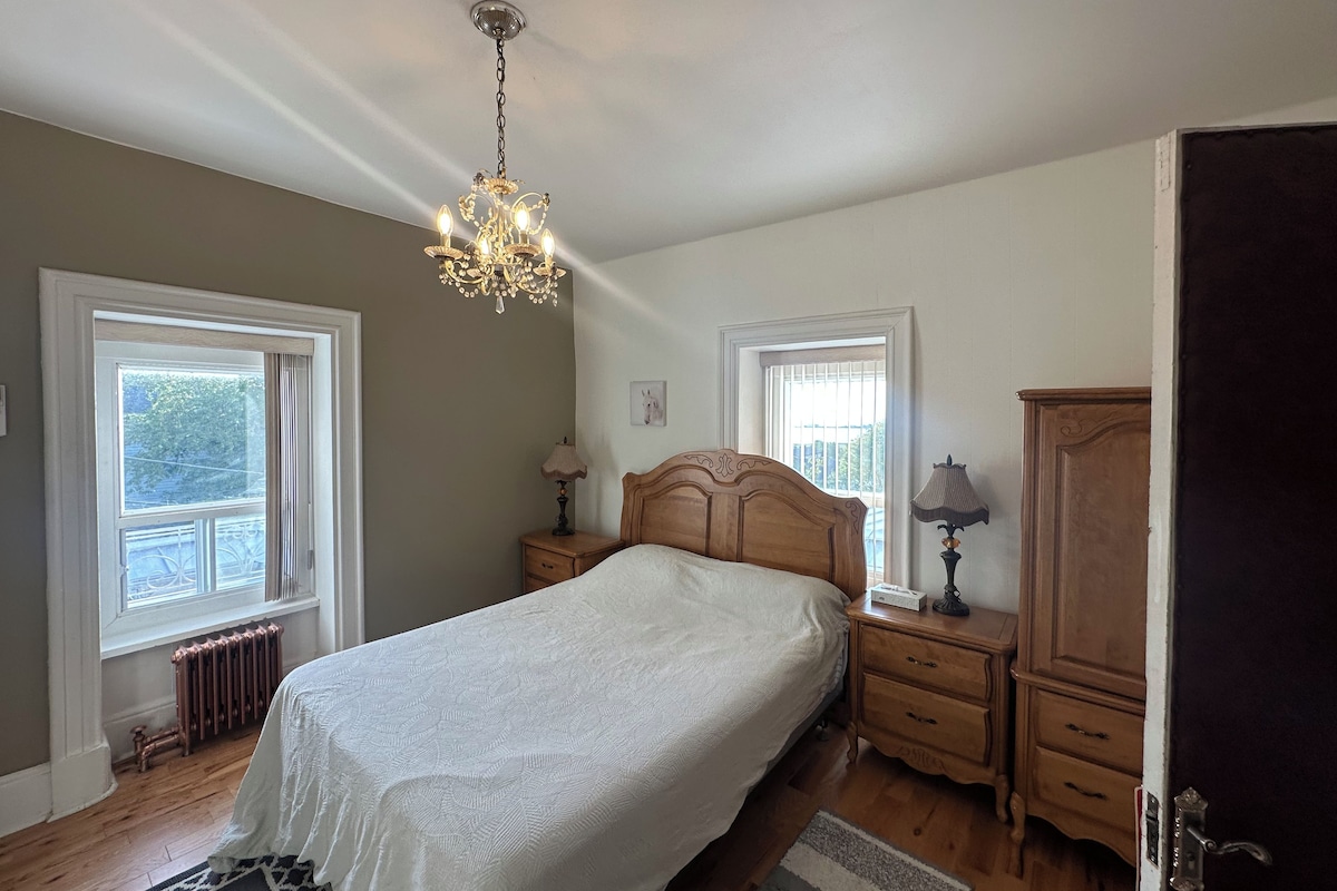 A bedroom with a bed, two bedside tables, a nightstand, a wooden wardrobe, a chandelier, and two windows with blinds.