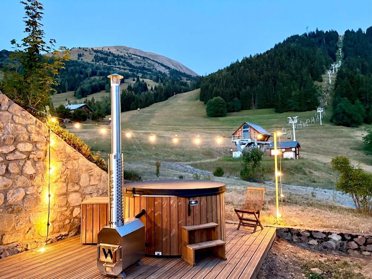 A hot tub on a wooden deck with a stone wall, surrounded by a mountainous landscape and a small cabin.