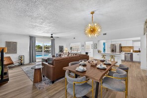 A modern living room with a brown leather sofa, a dining table set with plates and cups, and a kitchen area with white cabinets and stainless steel appliances. - The '59 Flamingo | Pool, Hot Tub & Cold Plunge (Apollo Beach)