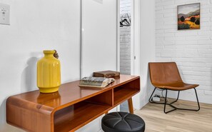 Ein Zimmer mit Schreibtisch aus Holz, einer gelben Vase, einem Buch, einem schwarzen Hocker, einem braunen Ledersessel und einer weißen Ziegelwand mit einem Gemälde.
