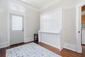 Ein Zimmer mit weißer Tür, einem weißen Kleiderschrank, einem kleinen Tisch und einem gemusterten Teppich auf Holzdielen.