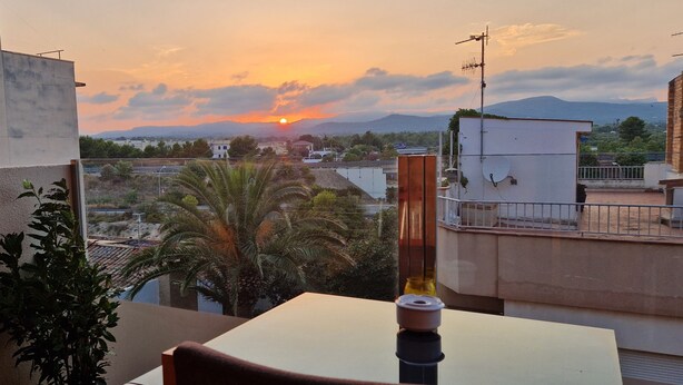 Ein Balkon mit Tisch und Stühlen, der auf einen Stadtblick mit Gebäuden und einem Sonnenuntergang blickt.
