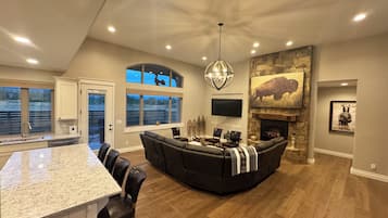 A spacious living area with a large black sectional sofa, a dining table with chairs, a kitchen area with a granite countertop, and a fireplace with a stone surround.