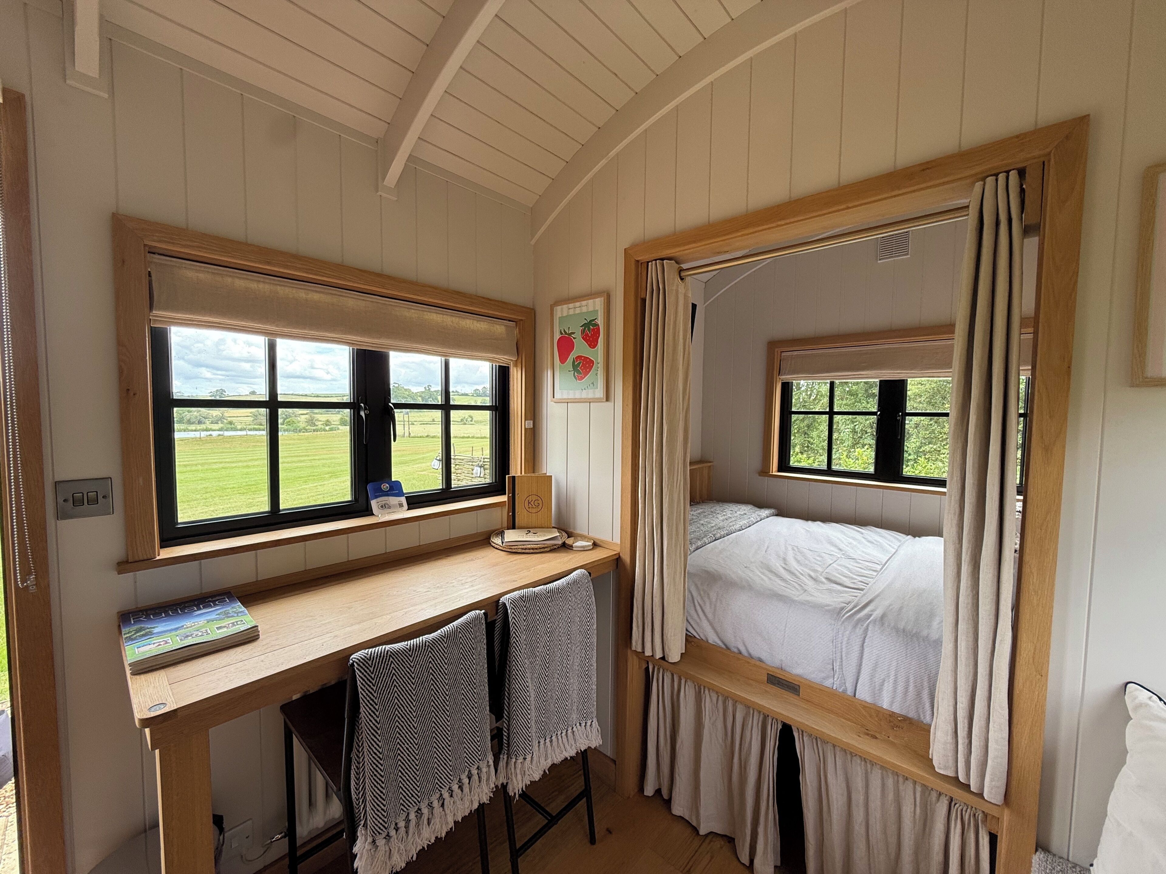 A room with a wooden desk, a bed with curtains, a window with a view of a green field, and a framed picture on the wall.