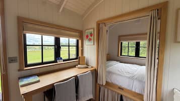A room with a wooden desk, a bed with curtains, a window with a view of a green field, and a framed picture on the wall.