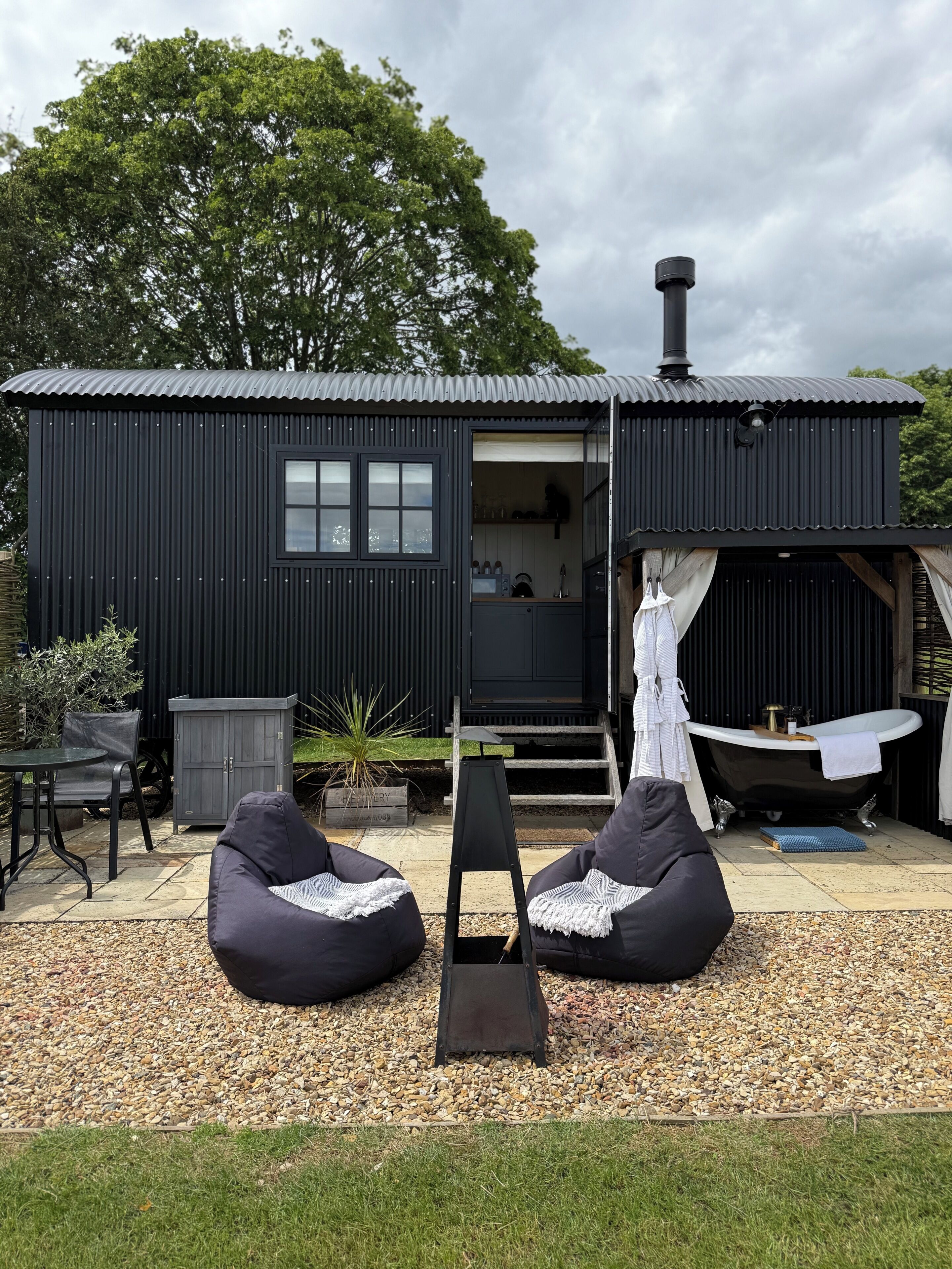 A black metal building with a small window and a door, a black bathtub, and two bean bag chairs on a gravel area.
