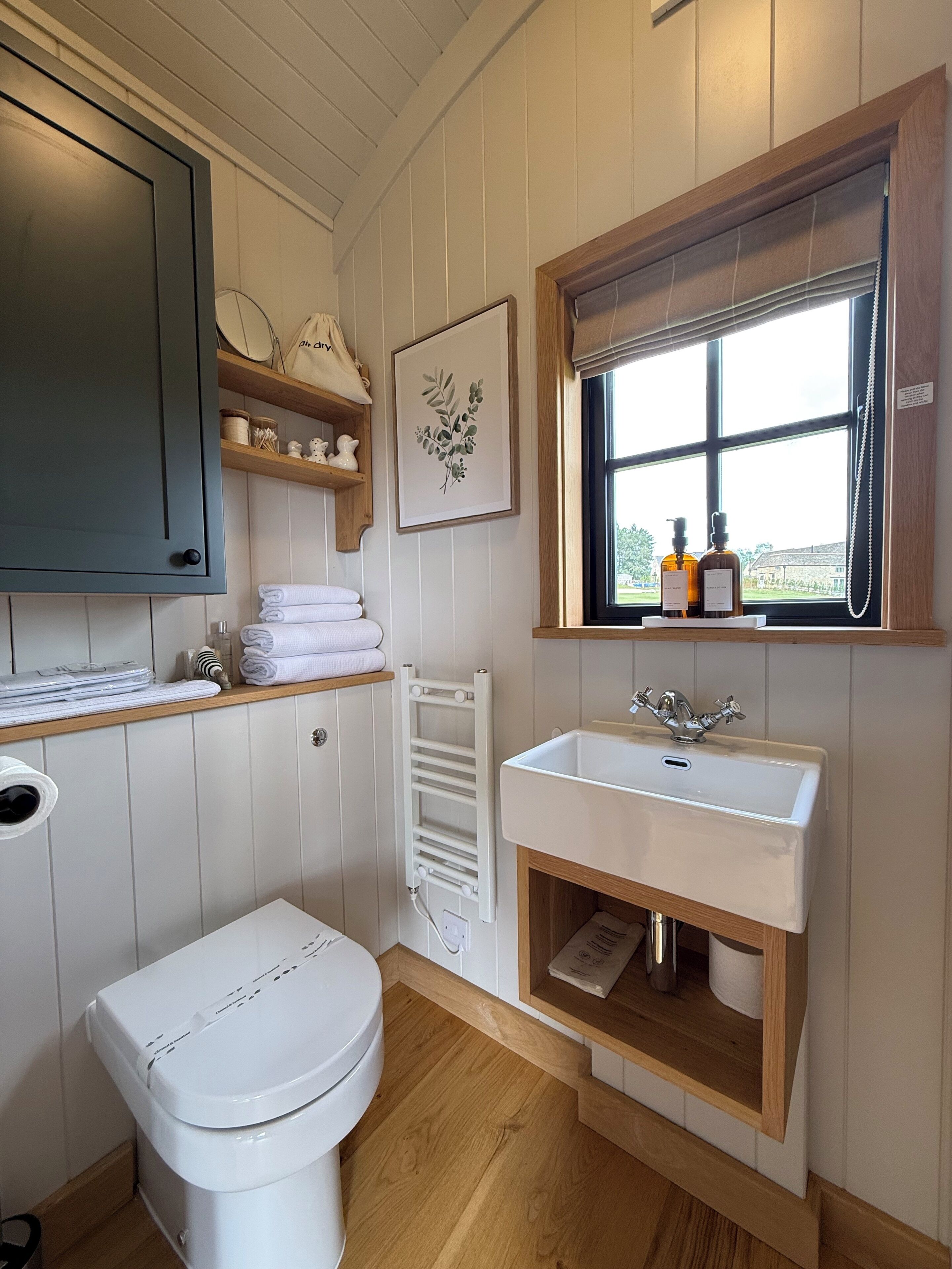 A bathroom with a toilet, a sink, a window, and a shelf with towels.