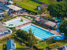Una gran piscina al aire libre con tobogán y área de juegos acuáticos, rodeada de sillones de descanso y un pequeño edificio.