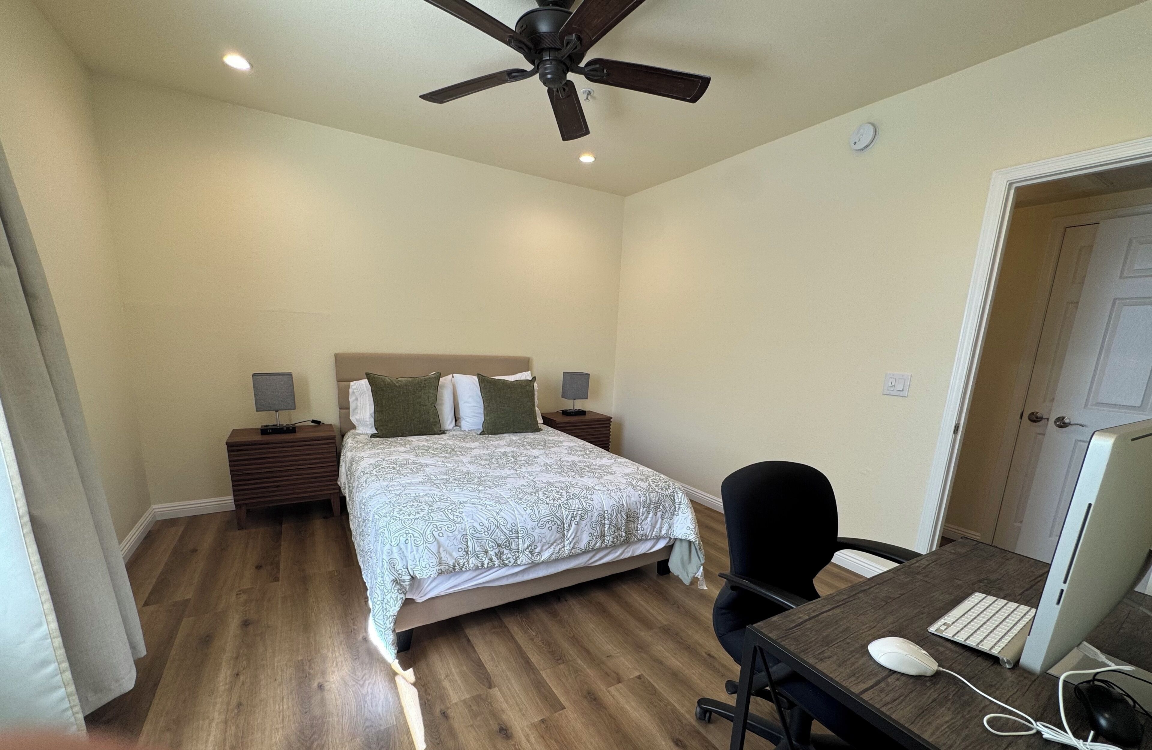 A bedroom with a bed, bedside tables, a desk with a chair, a computer, and a ceiling fan.