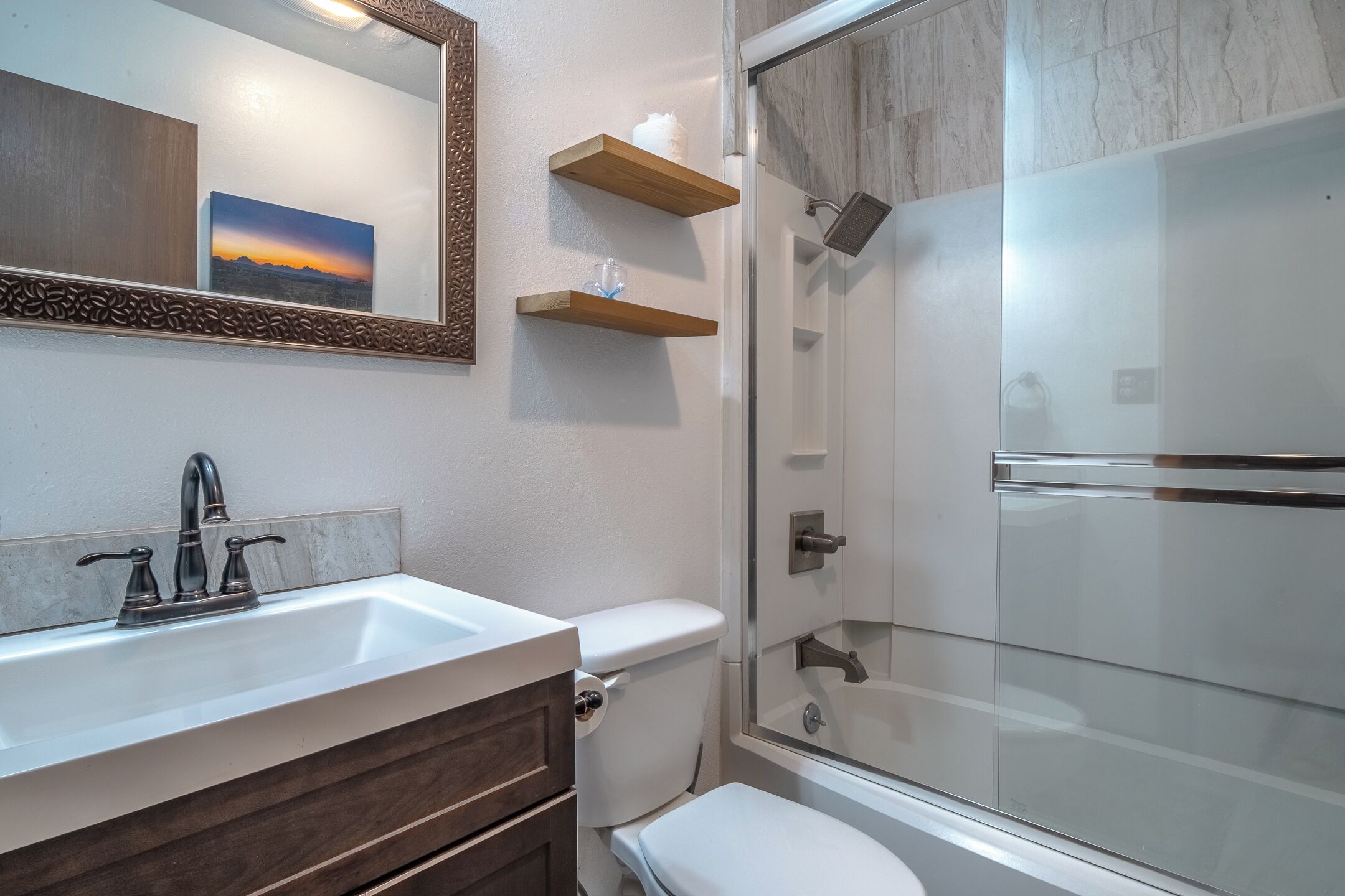 A bathroom with a white sink, a toilet, a shower with a glass door, and a wooden shelf above the toilet.