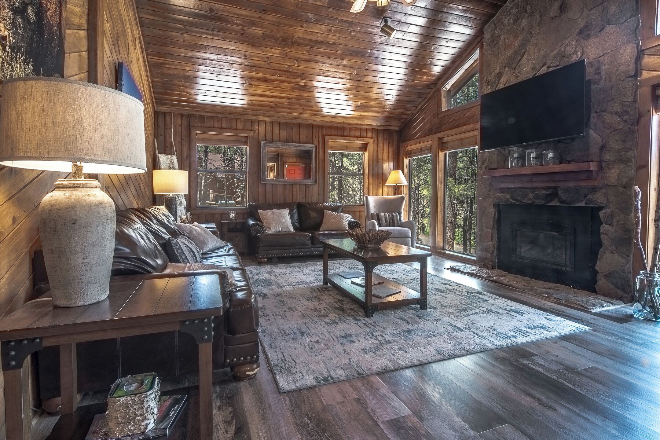 A log cabin interior with a fireplace, wooden furniture, and large windows.