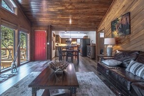 Een ruime woonkamer met houten plafond, een eettafel met stoelen, een leren bank en een keuken op de achtergrond.