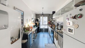 Una cocina estrecha con una isla central, un refrigerador adornado con imanes y fotografías, y un ventanal que proporciona luz natural.