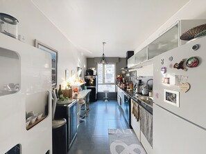 Eine schmale Küche mit einer zentralen Kochinsel, einem Kühlschrank mit Magneten und Fotos, und einem Fenster, das natürliches Licht hereinlässt.