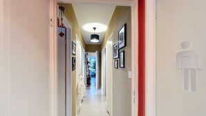 Un pasillo con piso blanco, paredes color beige y un marco de puerta rojo. Hay cuadros en las paredes y una lámpara en el techo.