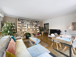 Ein Wohnzimmer mit einer großen Bücherwand, einem Weihnachtsbaum, einem Esstisch mit Stühlen und einem Sofa mit Kissen.