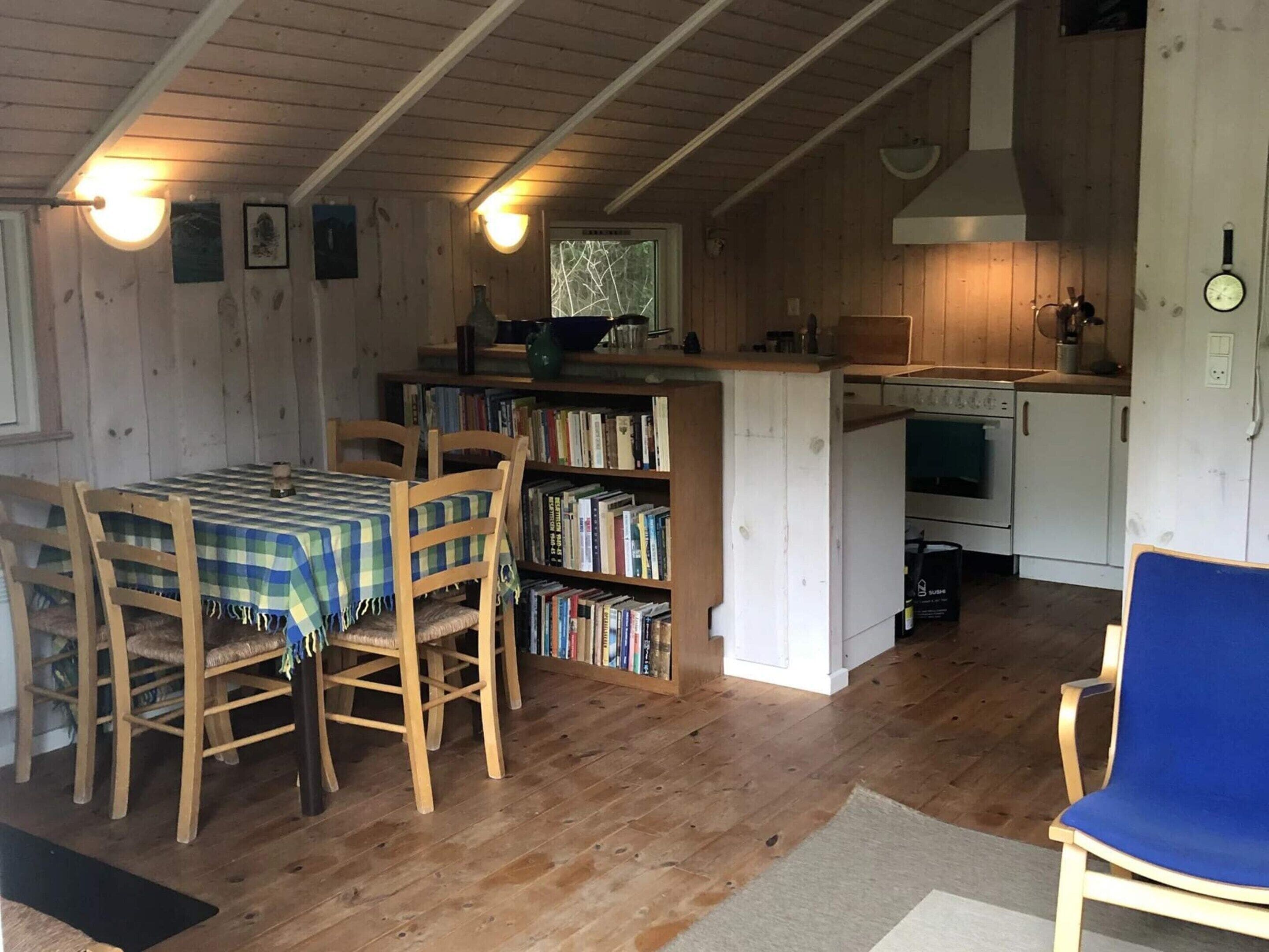 A cozy cabin interior with a dining area, a bookshelf, a kitchen with a stove, and a clock on the wall.