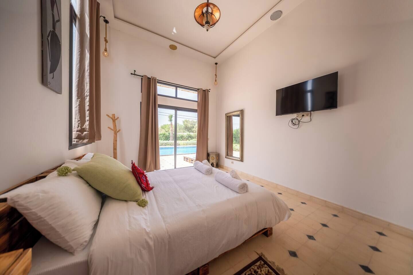 A bedroom with a large bed, a flat-screen TV mounted on the wall, and a view of a pool through the window.