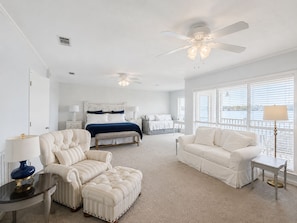 A bedroom with a bed, two bedside tables, a nightstand, a chair, an ottoman, a sofa, and a window with blinds.