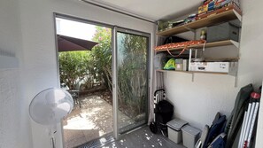 Ein Zimmer mit einer Schiebetür zu einem Garten, einem kleinen Tisch mit einem Stuhl, einem Regal mit Gesellschaftsspielen und einer Mülltonne.