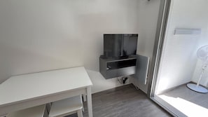 Ein kleiner Raum mit einem weißen Schreibtisch, einem Wandfernseher und einem Standventilator.