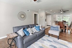 A modern living room with a grey sofa, a small wooden side table, a patterned rug, and a dining area with a wooden table and chairs. - Hot tub, Game room, Pool, 7 miles to stockyards (Haltom City)