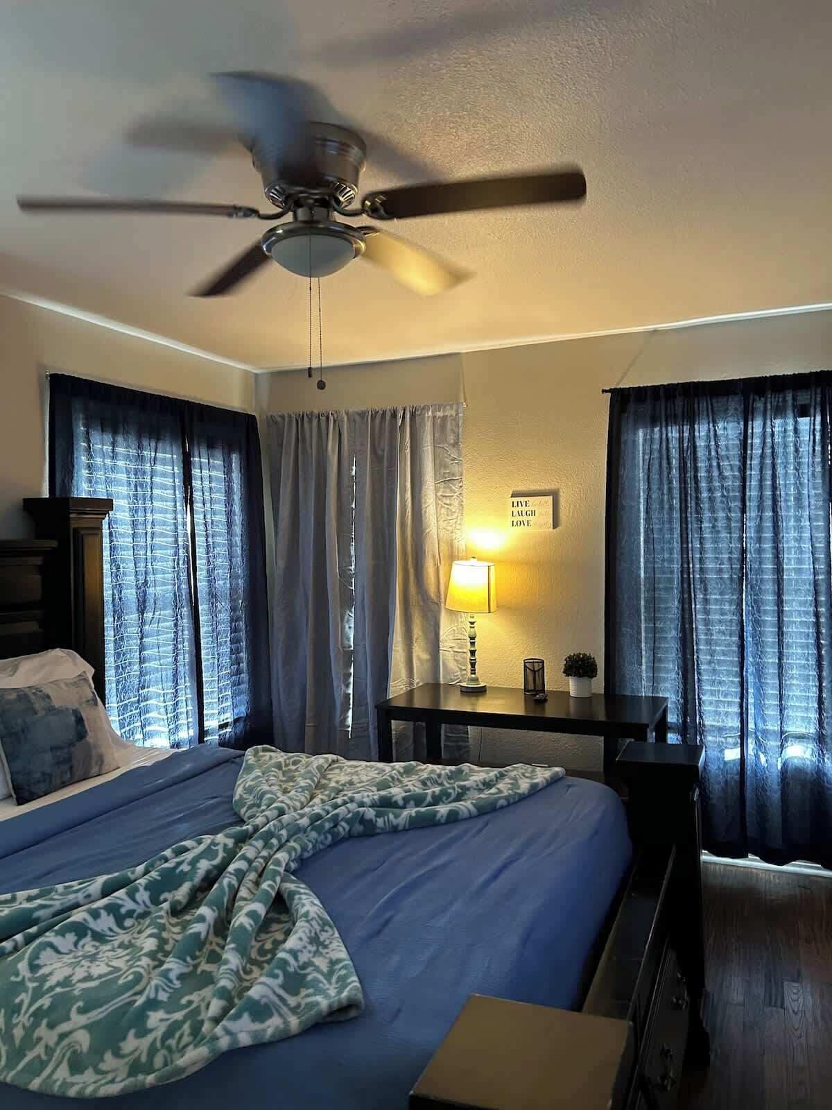 A hotel room with a bed, a desk, a ceiling fan, and curtains.