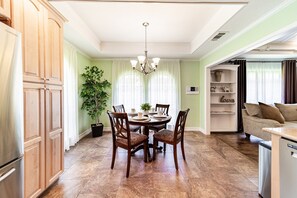 Ein Essbereich mit einem runden Tisch, Stühlen und einer Chandelieber. Eine Kübelpflanze und ein Regal mit Dekorationsobjekten sind ebenfalls vorhanden.