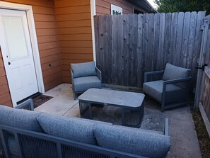 En terrasse med et træhegn, en hvid dør og grå udendørs møbler.