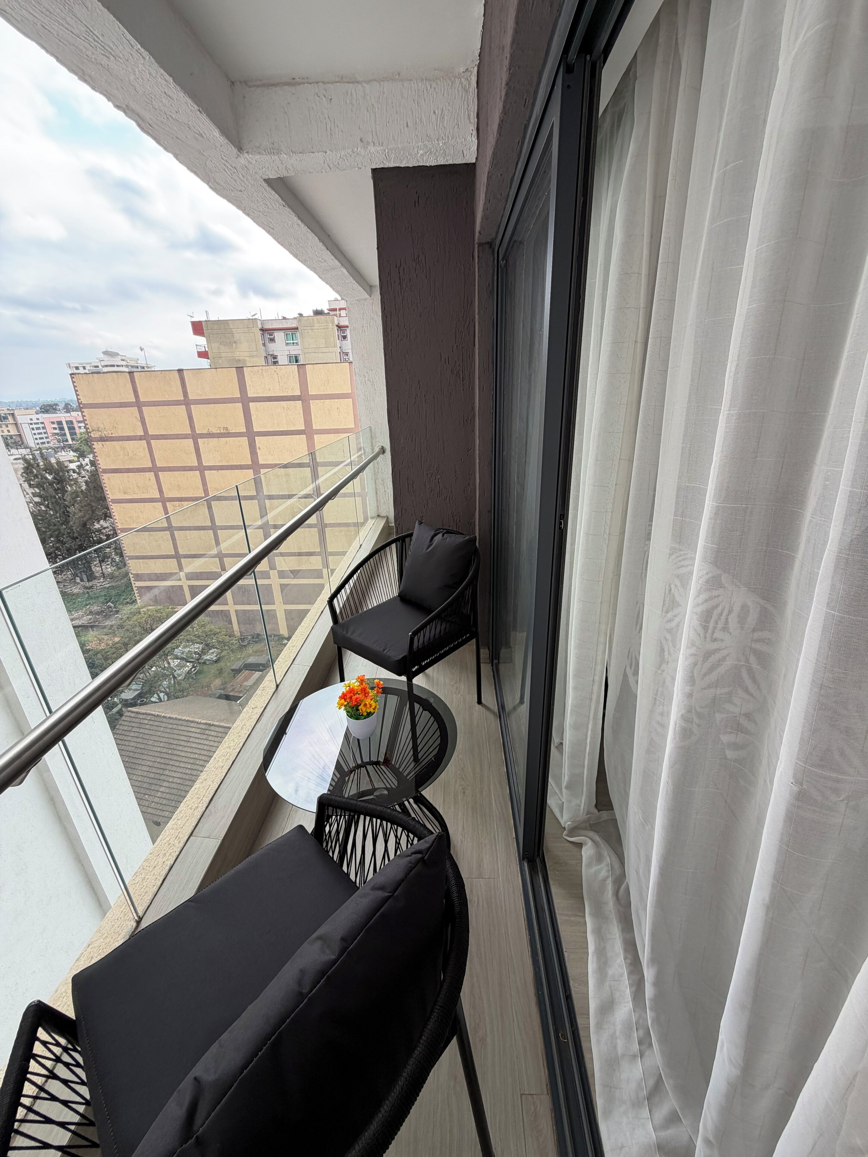 Een balkon met twee zwarte stoelen, een kleine tafel met een bloevas, en uitzicht op gebouwen.