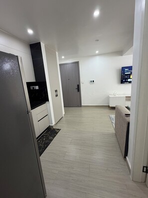 A modern kitchen with a refrigerator, cabinets, and a microwave. There is a door leading to another room, a television mounted on the wall, and a small white table with a vase. - Cozy & Stylish Apartment with stunning views, 2 minutes from yaya center. (Nairobi)