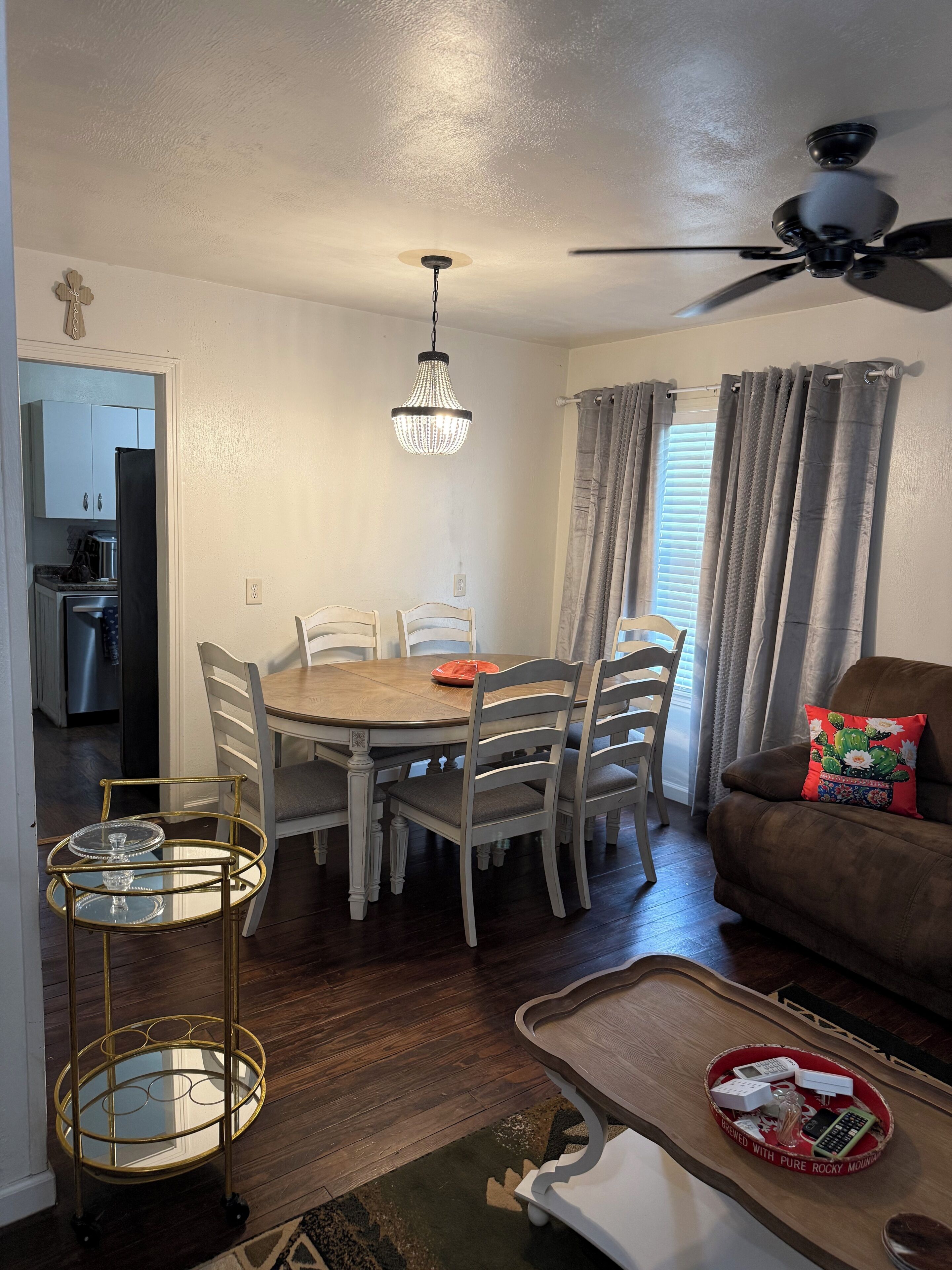 A dining area with a chandelier, a round table, and chairs. There is a kitchen visible in the background.