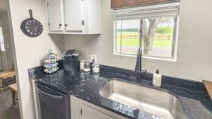 A kitchen with white cabinets, a black countertop, a coffee maker, a clock, and a window with a view of a green landscape. - Brittleville Bunkhouse: Peaceful Country Getaway (Brooksville)
