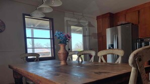 A kitchen with a wooden table, chairs, a vase with flowers, a refrigerator, and a window with a view.