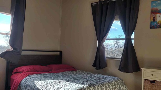 A bedroom with a bed, a window with curtains, a painting on the wall, and a small cabinet.