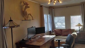A room with a wooden desk, a computer, a chair, a sofa, a decorative deer wall piece, and a window with curtains.